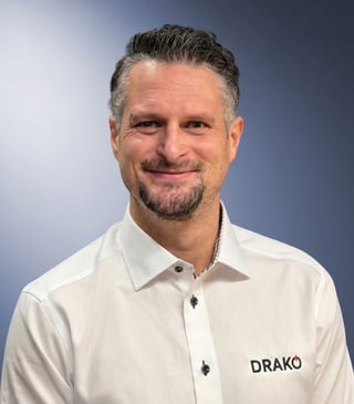 Professional headshot of a middle-aged man with gray hair and beard wearing a white DRAKO polo shirt against a blue gradient background
