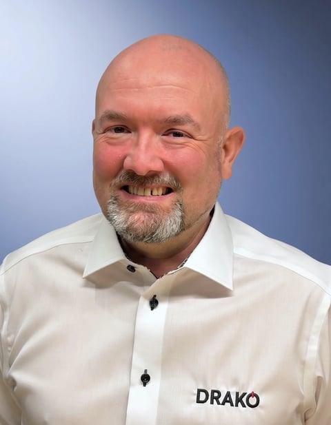 Professional headshot of a man with light skin, bald head, and gray beard wearing a white shirt with DRAKO branding against a blue background
